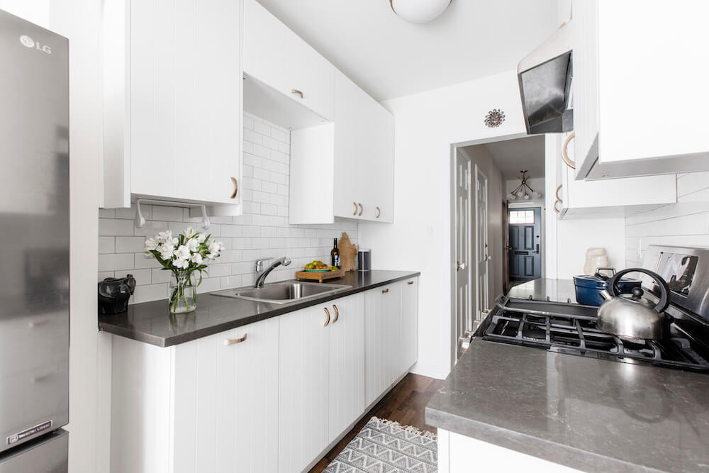 black granite countertop in a white galley kitchen with white kitchen cabinets with handles and white backsplash after renovation