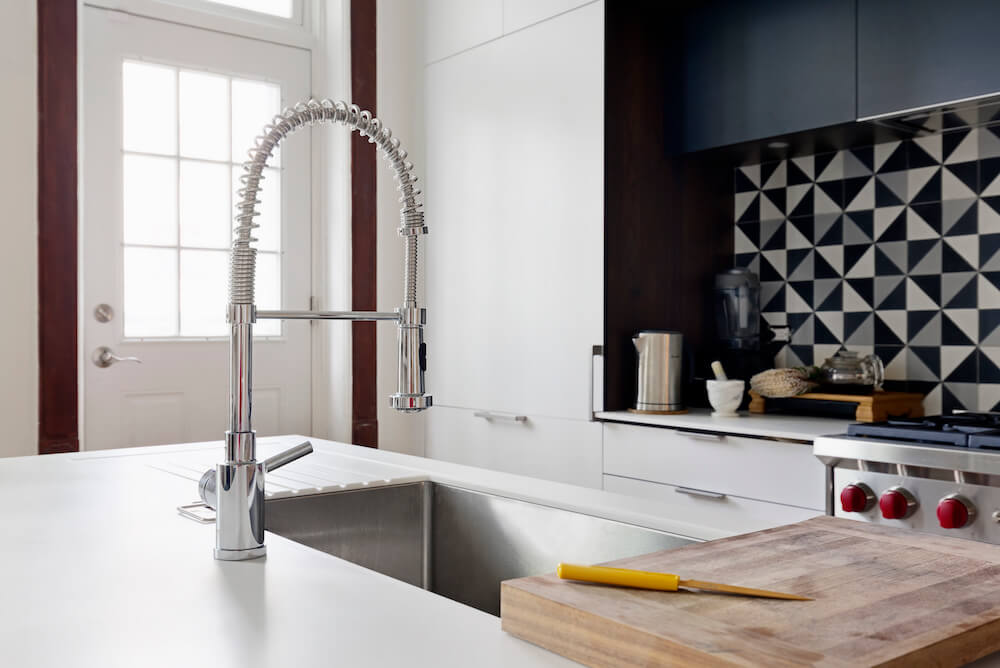 White kitchen island with undermount sink and multifunctional faucet facing kitchen with abstract backsplash after renovation