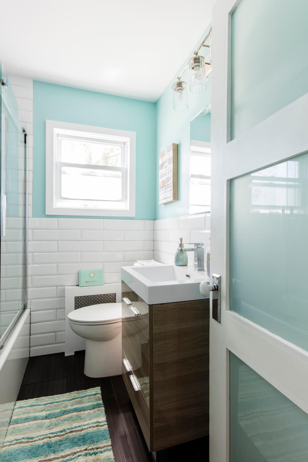 White subway tiles on half wall with glossy brown vanity and white sink and brown wooden floor tiles after renovation